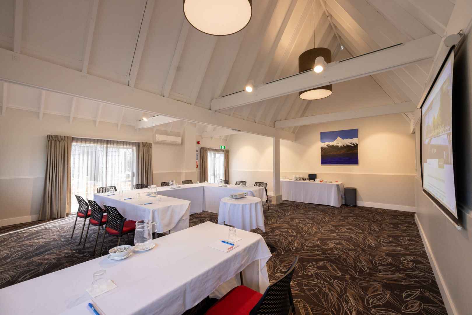 White-plastered function room with vaulted timber-truss ceiling, white tablecloths, projection screen, and patterned carpet