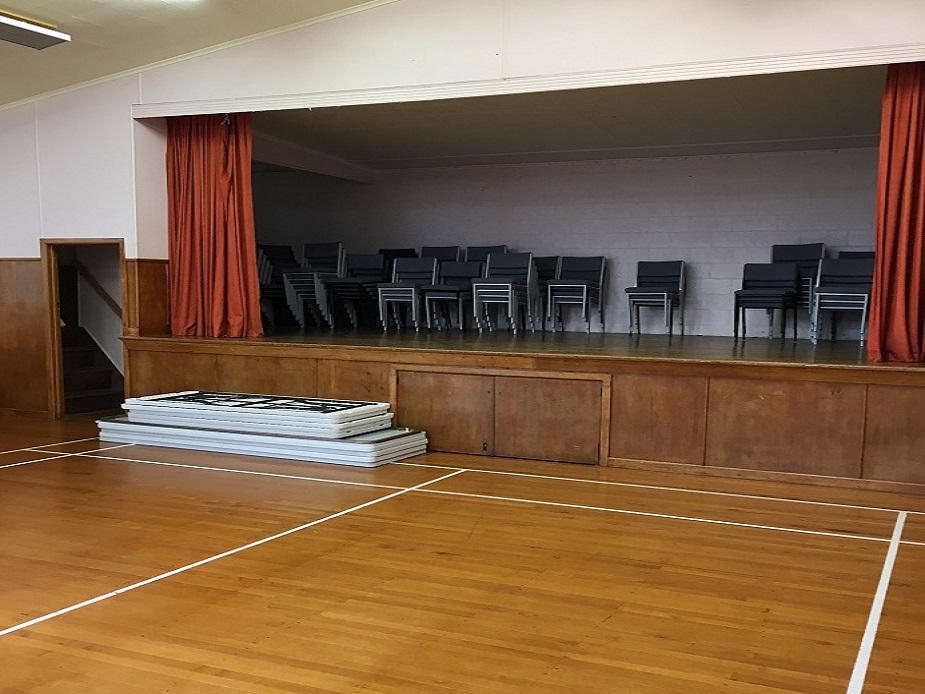Hall interior with timber floor, raised timber platform with red curtains, and stacked chairs