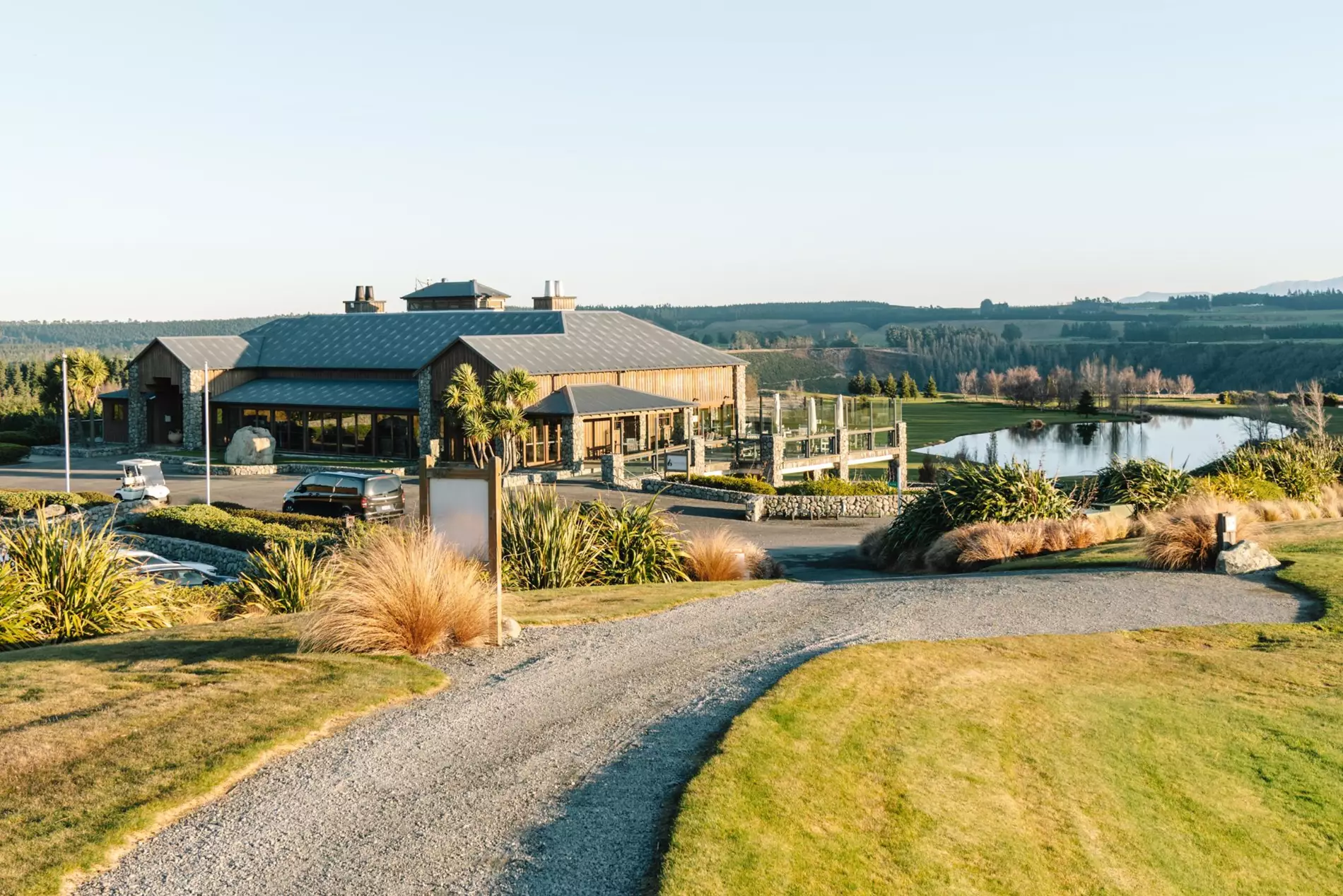 Rural resort building with metal roof beside a pond, viewed from a gravel driveway