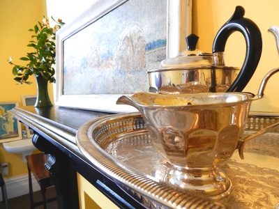 Detail of a silver tea service on a tray with a painting and flowers on a mantelpiece behind