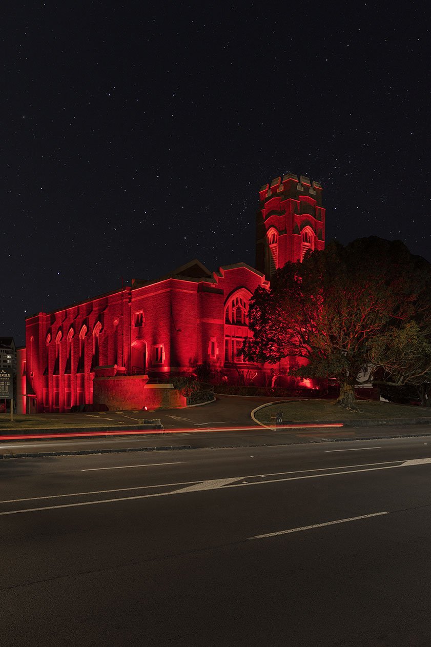 Red brick church with castellated tower, illuminated in red at night, viewed from across the road