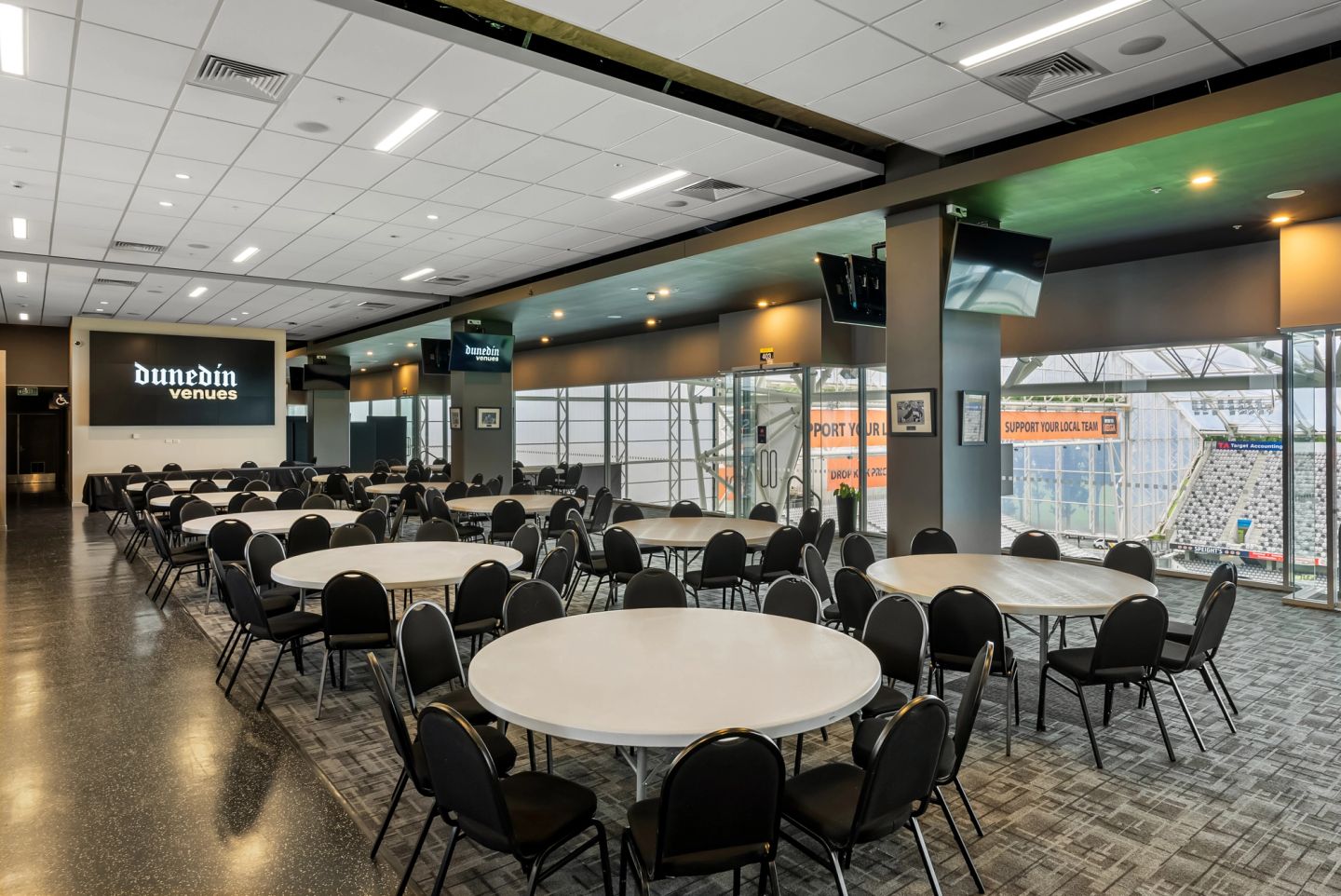 Stadium function room with round white tables, black chairs, wall-mounted screens, glass wall with field and stands beyond