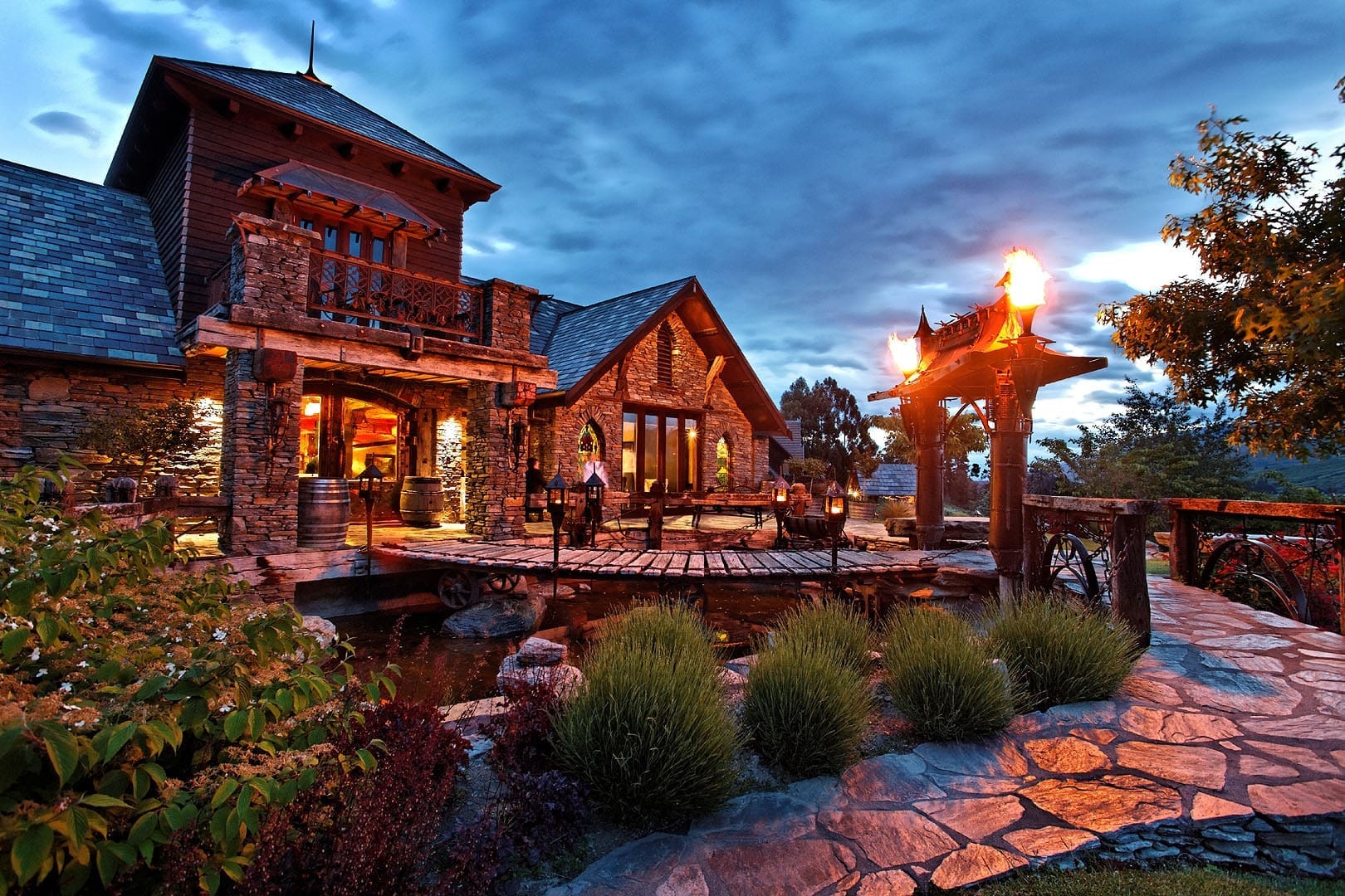 Stone and timber estate building at dusk with lit torch flares, pond, and garden terrace