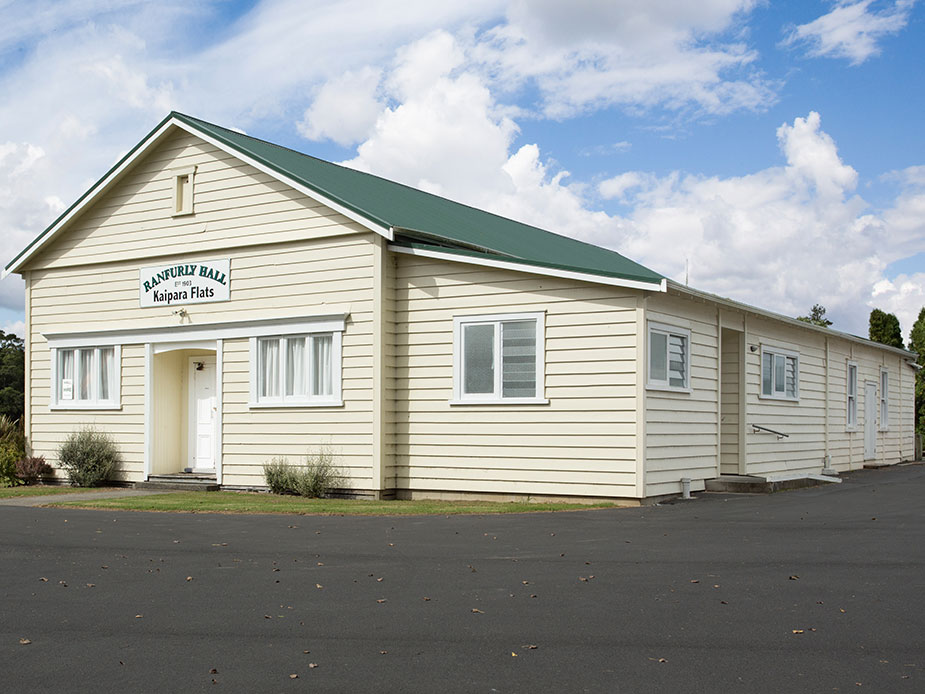 Cream weatherboard hall with green metal roof, gabled entry, and car park frontage