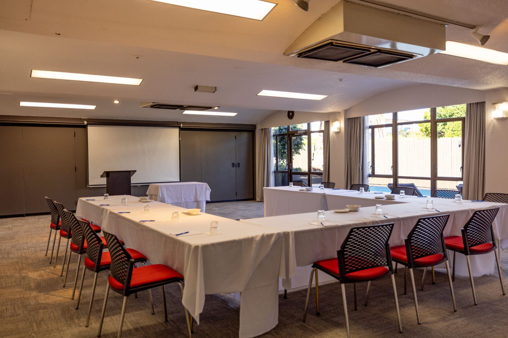Plain conference room with white tables in U-shape layout, red-seated chairs, and projection screen