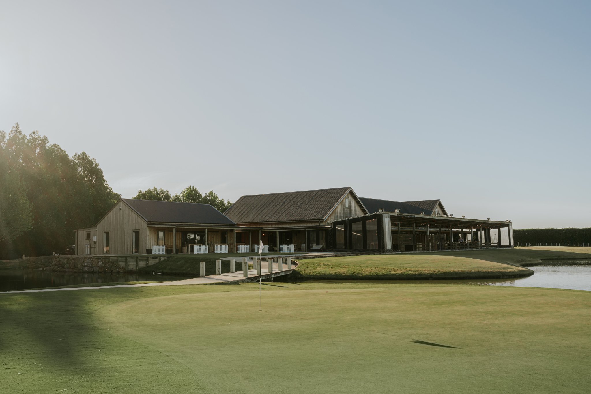 Timber and steel clubhouse beside a putting green with pond and trees, low afternoon light