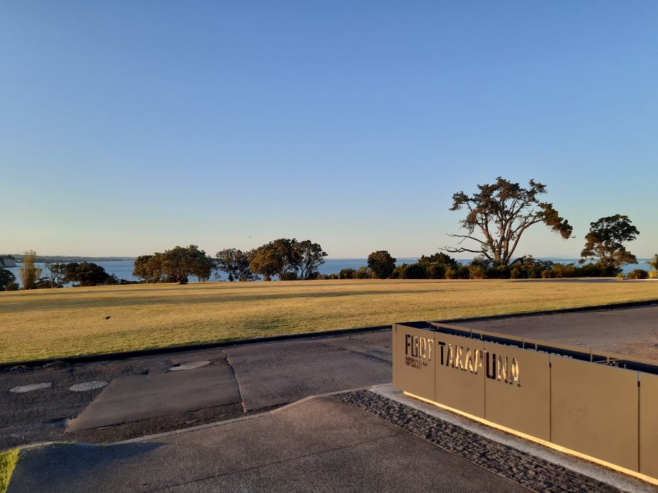 Open grassy headland with trees and coastal water views at golden hour, Fort Takapuna sign