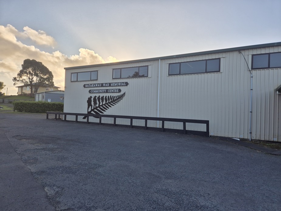 White corrugated-iron hall exterior with fern silhouette mural, car park in foreground