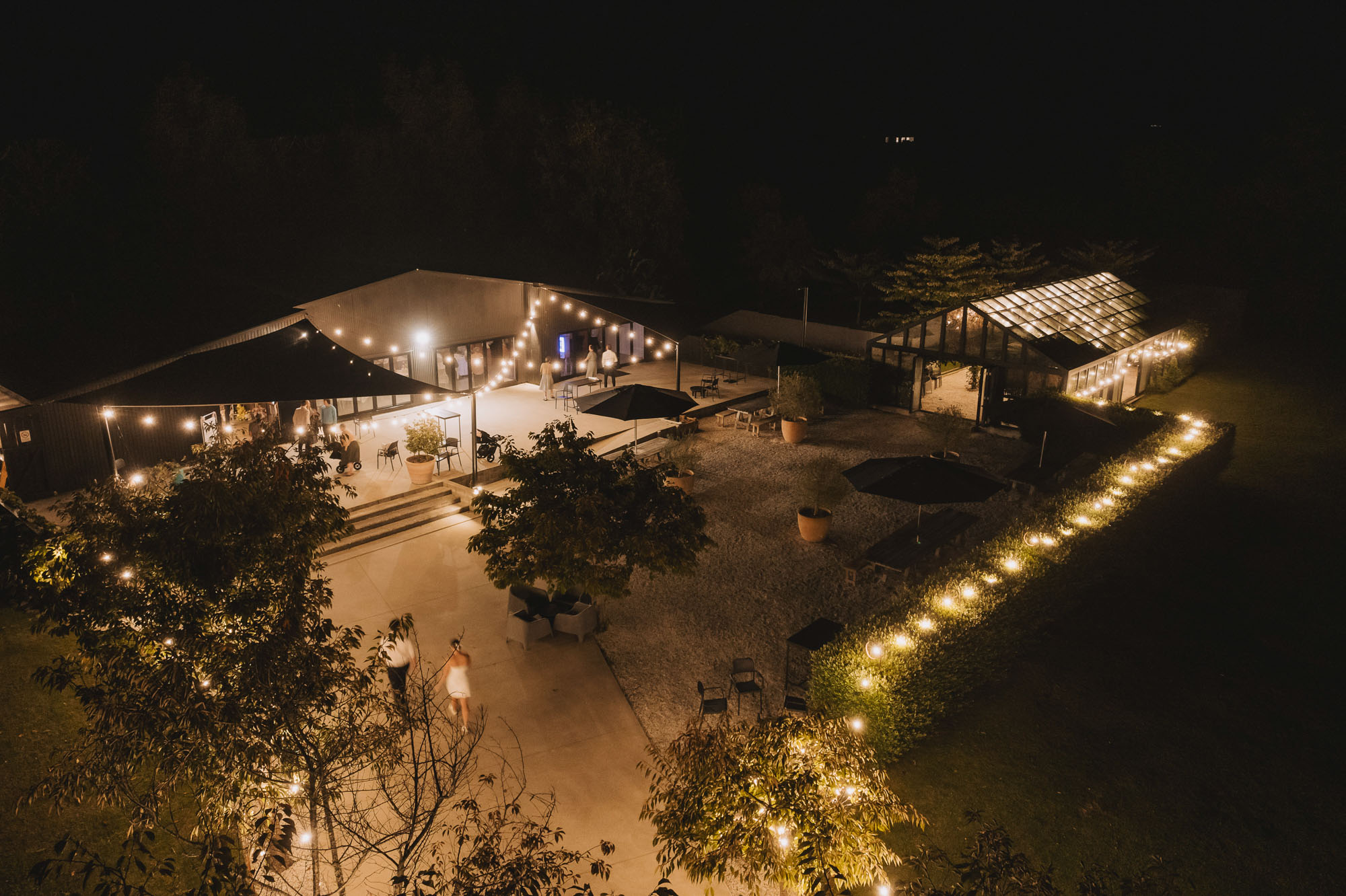 Night aerial of lit venue buildings and string lights on gravel courtyard