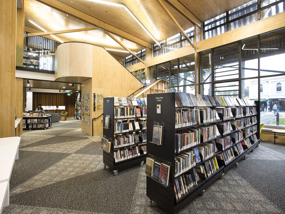 Open-plan library interior with timber ceiling, book shelving, mezzanine stair, and full-height glazed frontage