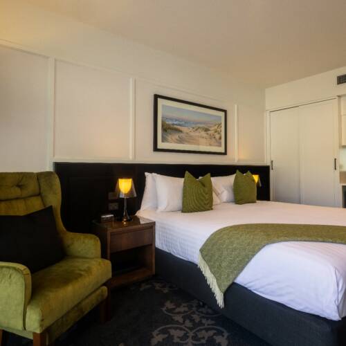 Hotel room with double bed, green velvet armchair, landscape artwork above headboard, warm lighting