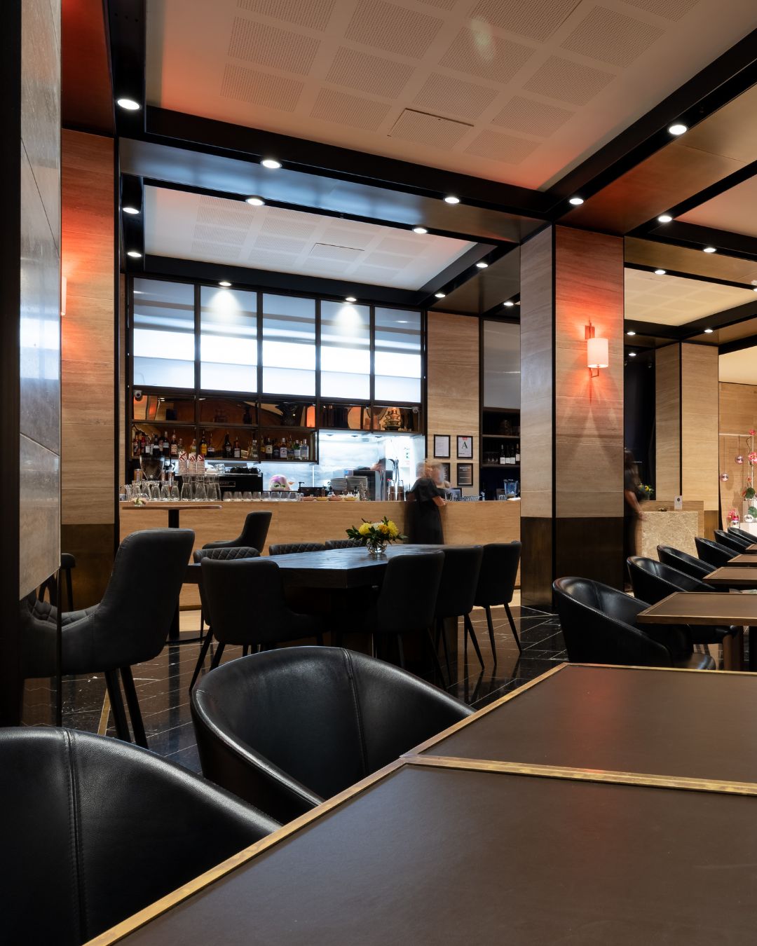 Dark-toned dining room with black leather chairs, round tables, wood-panelled walls, bar visible through opening