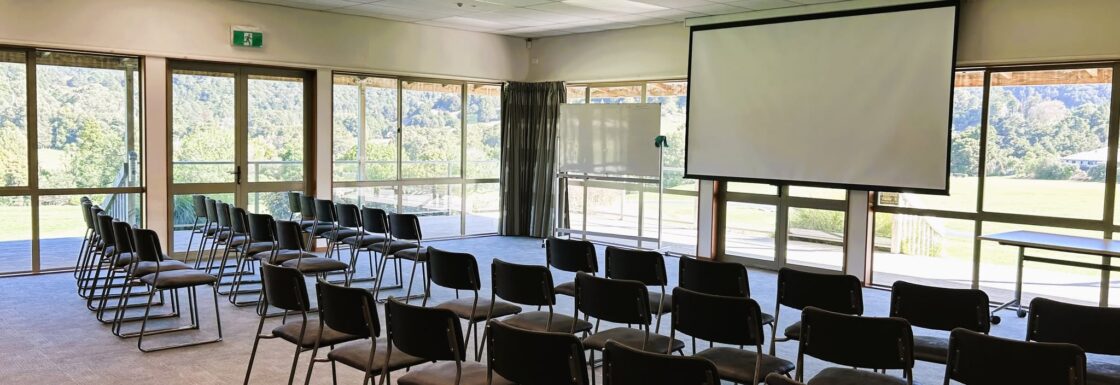 Meeting or function room with rows of chairs facing a projector screen, flanked by large windows overlooking greenery