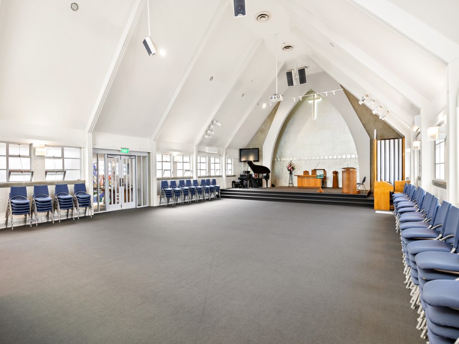 White-painted vaulted hall with grey carpet, chairs around the perimeter, and a raised platform at one end