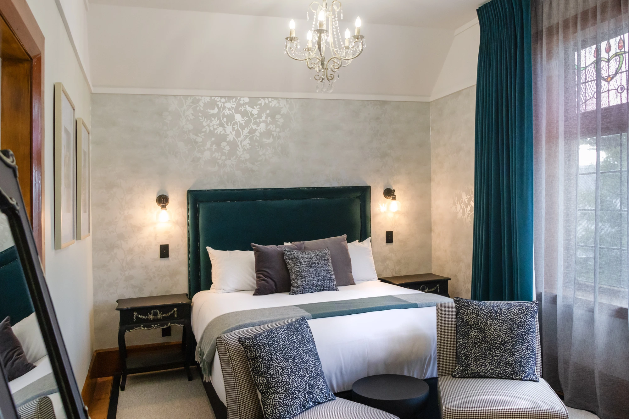 Guest room with dark green upholstered bed, crystal chandelier, patterned wallpaper, and stained-glass window