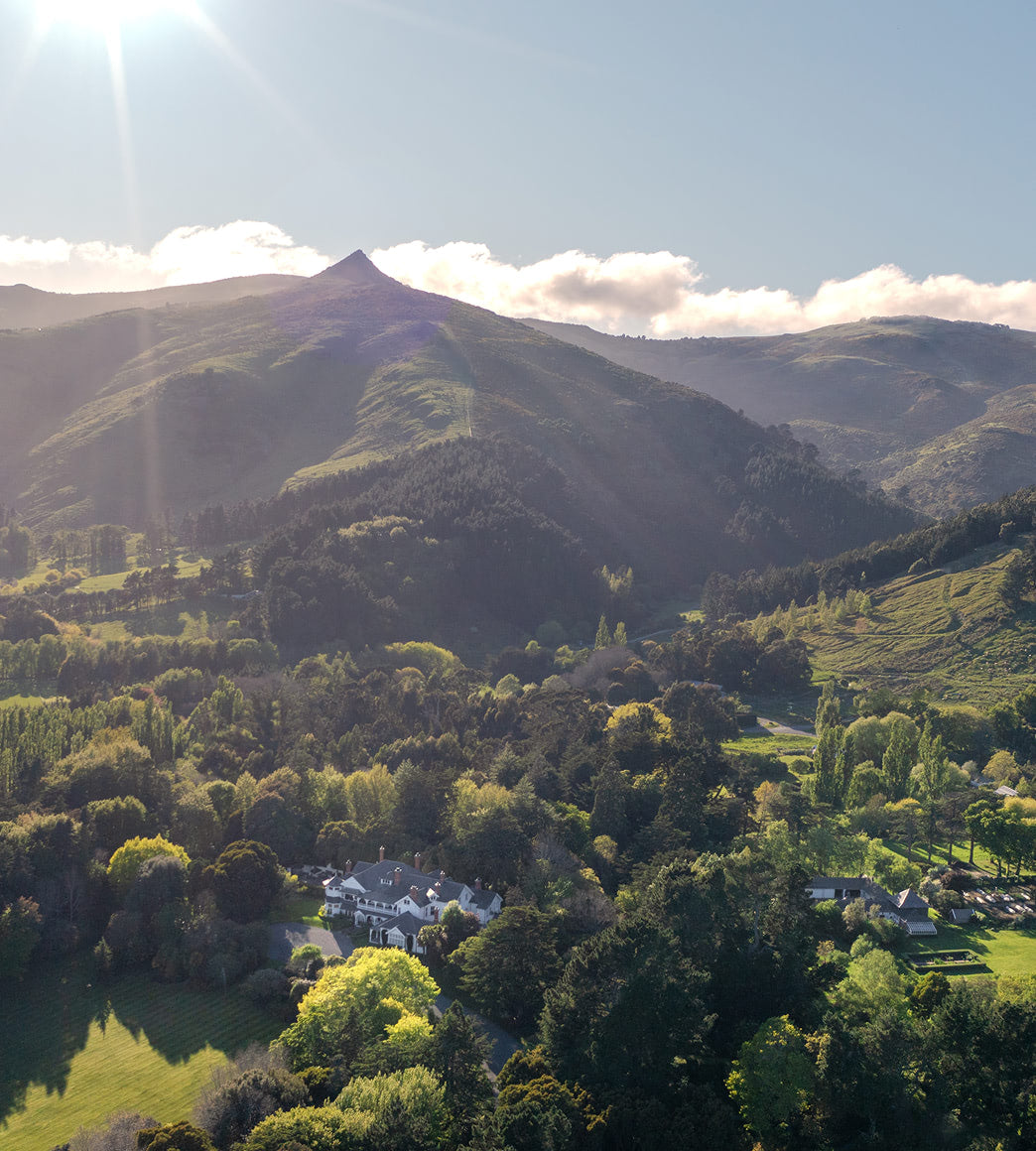 Aerial view of a white manor house and gardens set in a valley with forested hills and a pointed peak