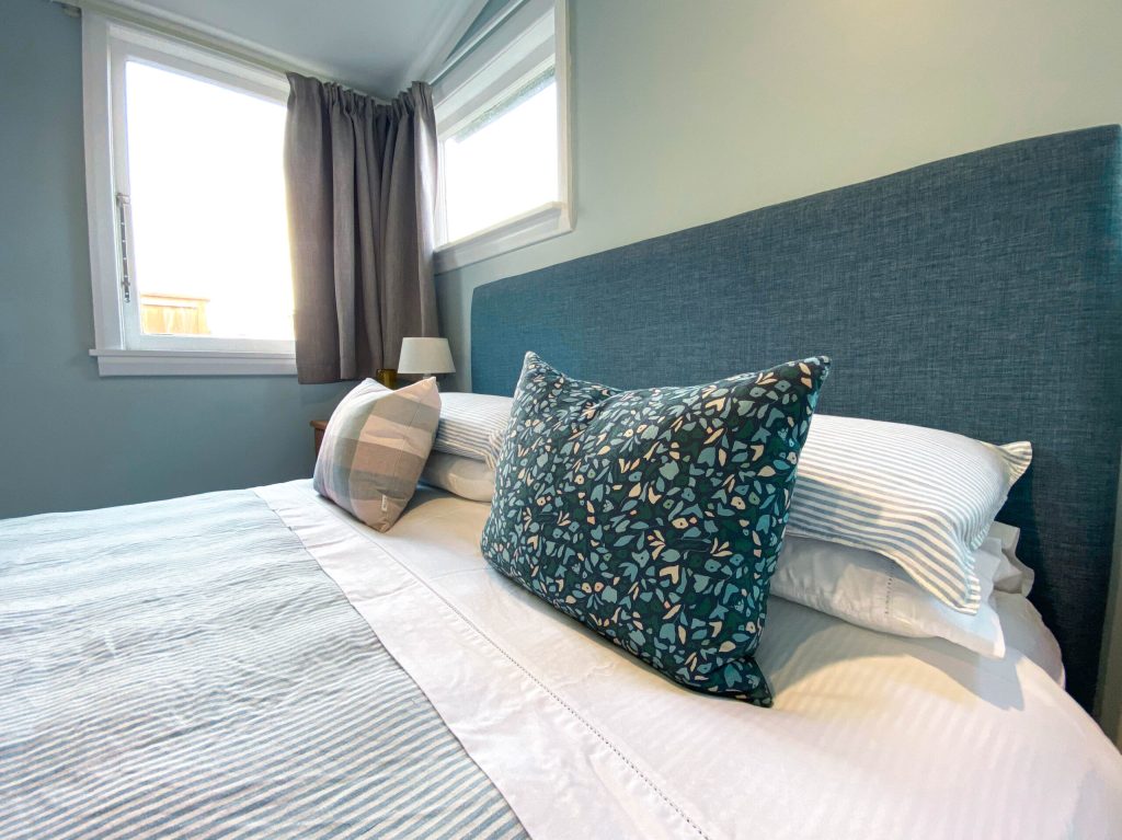 Blue-walled bedroom with upholstered headboard, layered cushions, and curtained window