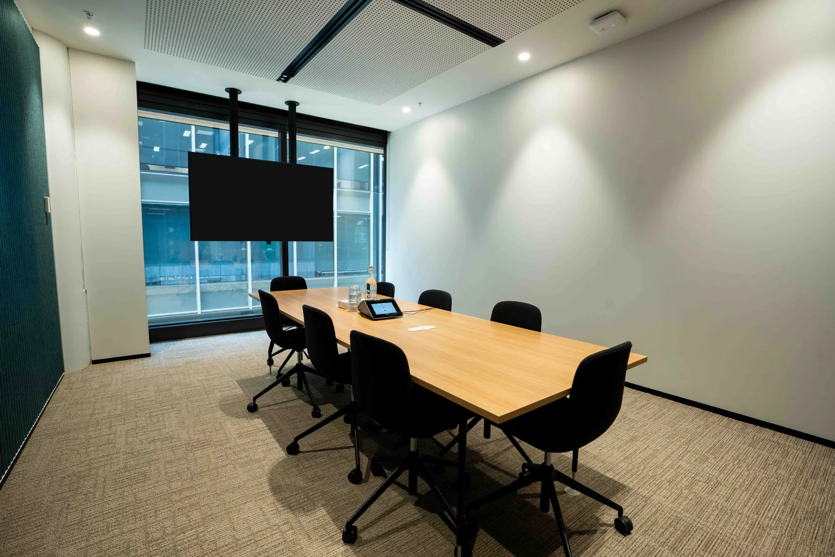 Meeting room with timber-topped table, black chairs, ceiling-mounted screen, and floor-to-ceiling city-view windows