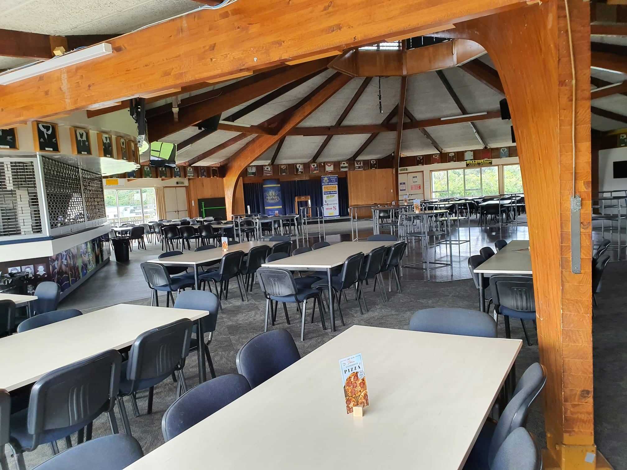 Large club room with circular timber ceiling, long tables and chairs, and a small raised platform at the far end