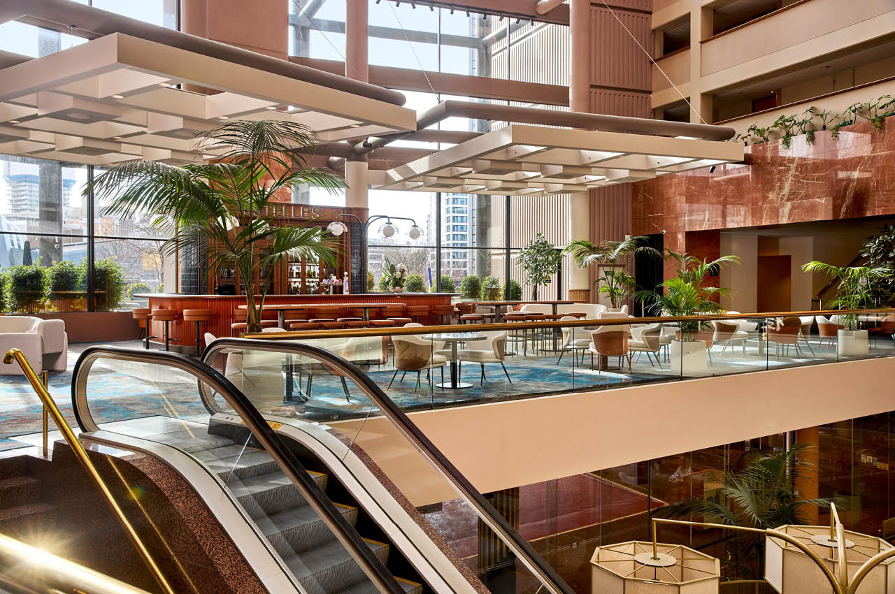 Multi-level atrium with escalators, indoor palms, pink stone walls, and a cafe bar below