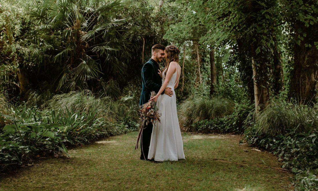 Two people embracing in a dense garden clearing surrounded by trees and lush planting