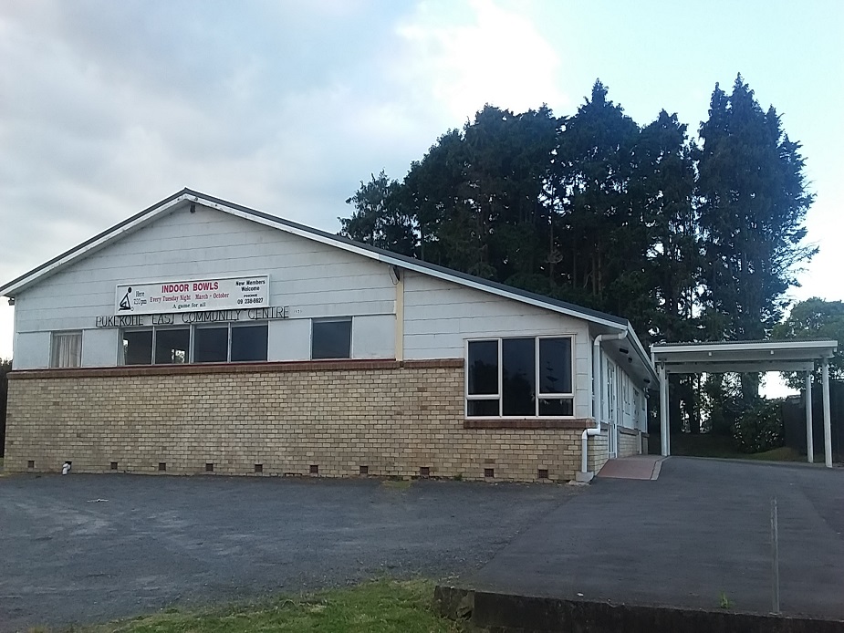 Single-storey white-painted brick community hall with low-pitched roof and car park, trees behind