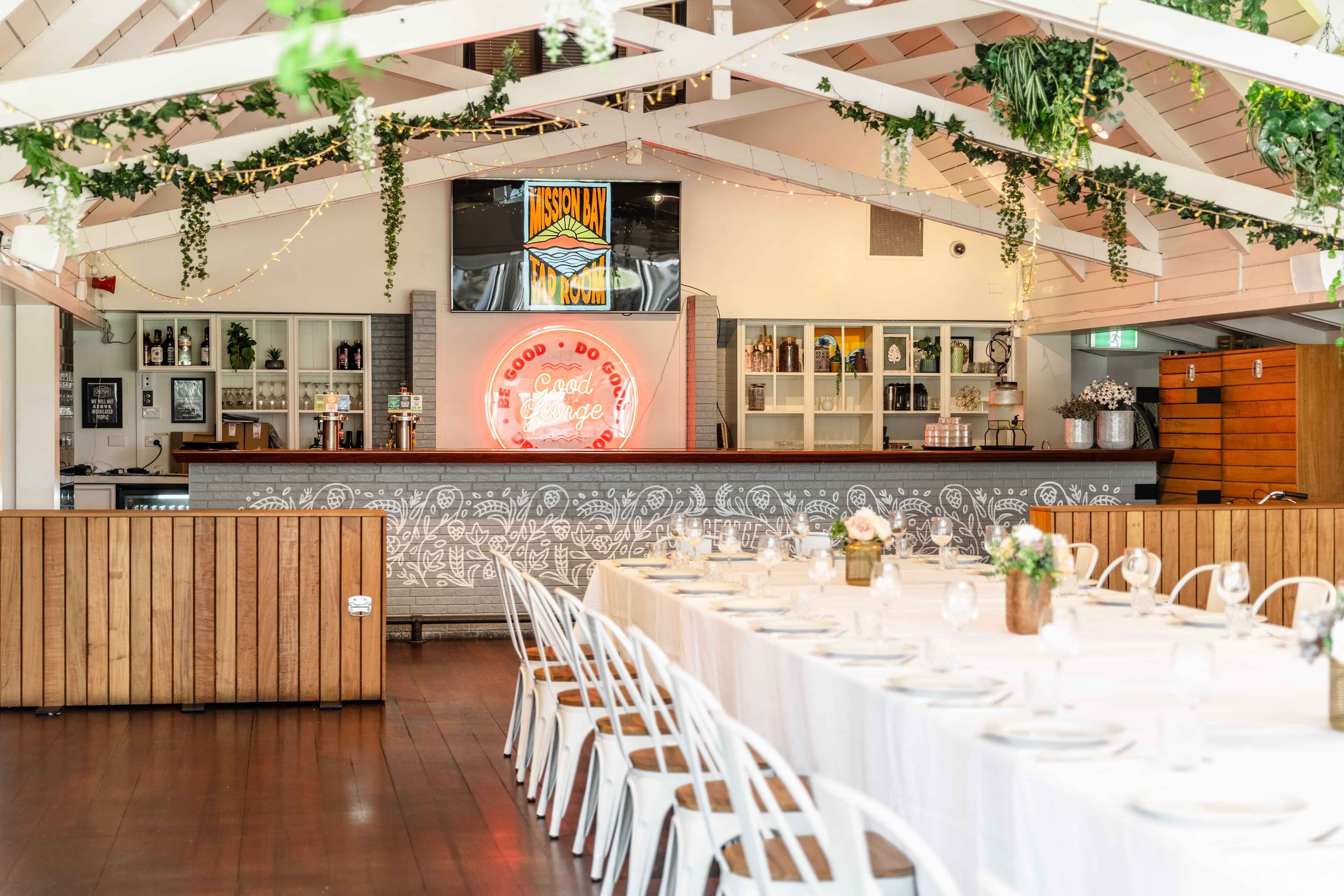 Function hall with vaulted white timber ceiling, neon bar sign and round tables set for dining