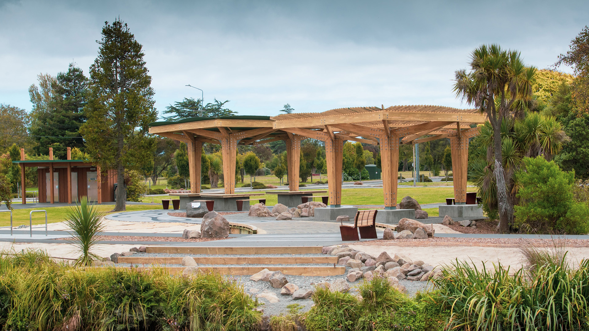 Timber open pavilion structure with latticed roof beams, concrete seating steps and boulders, parkland setting