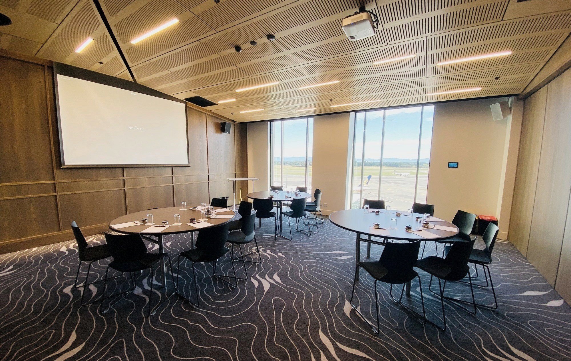 Timber-panelled meeting room with round tables, black chairs, projection screen, and large windows