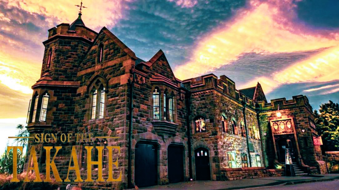 Gothic stone building with turrets, arched windows and a central tower, lit at dusk against a dramatic sky