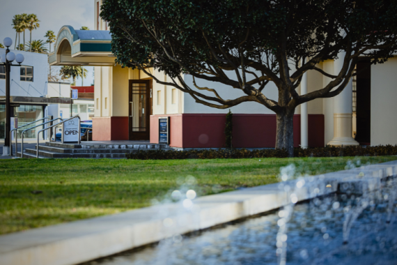 Art deco theatre exterior of cream and red render with a bare tree and water feature, street-level view