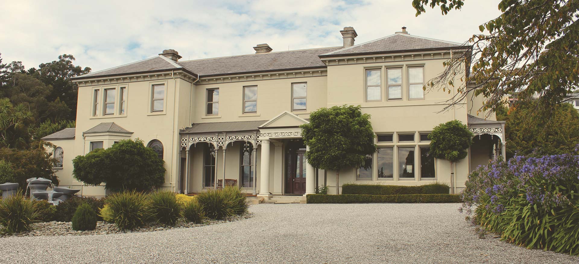 Two-storey cream Victorian manor with verandah and arched windows behind a gravel driveway and clipped hedges