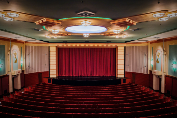 Art deco auditorium with tiered red seating, circular ceiling medallion, wall sconces, and a red curtained stage