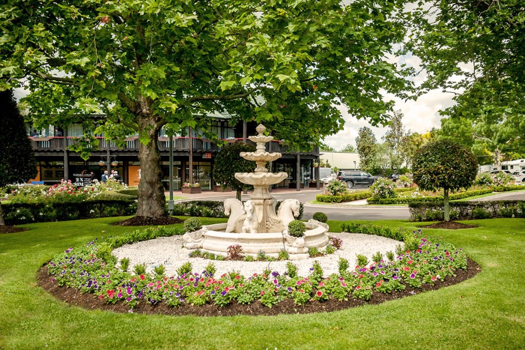 Ornate stone tiered fountain surrounded by a circular flower bed and mature trees
