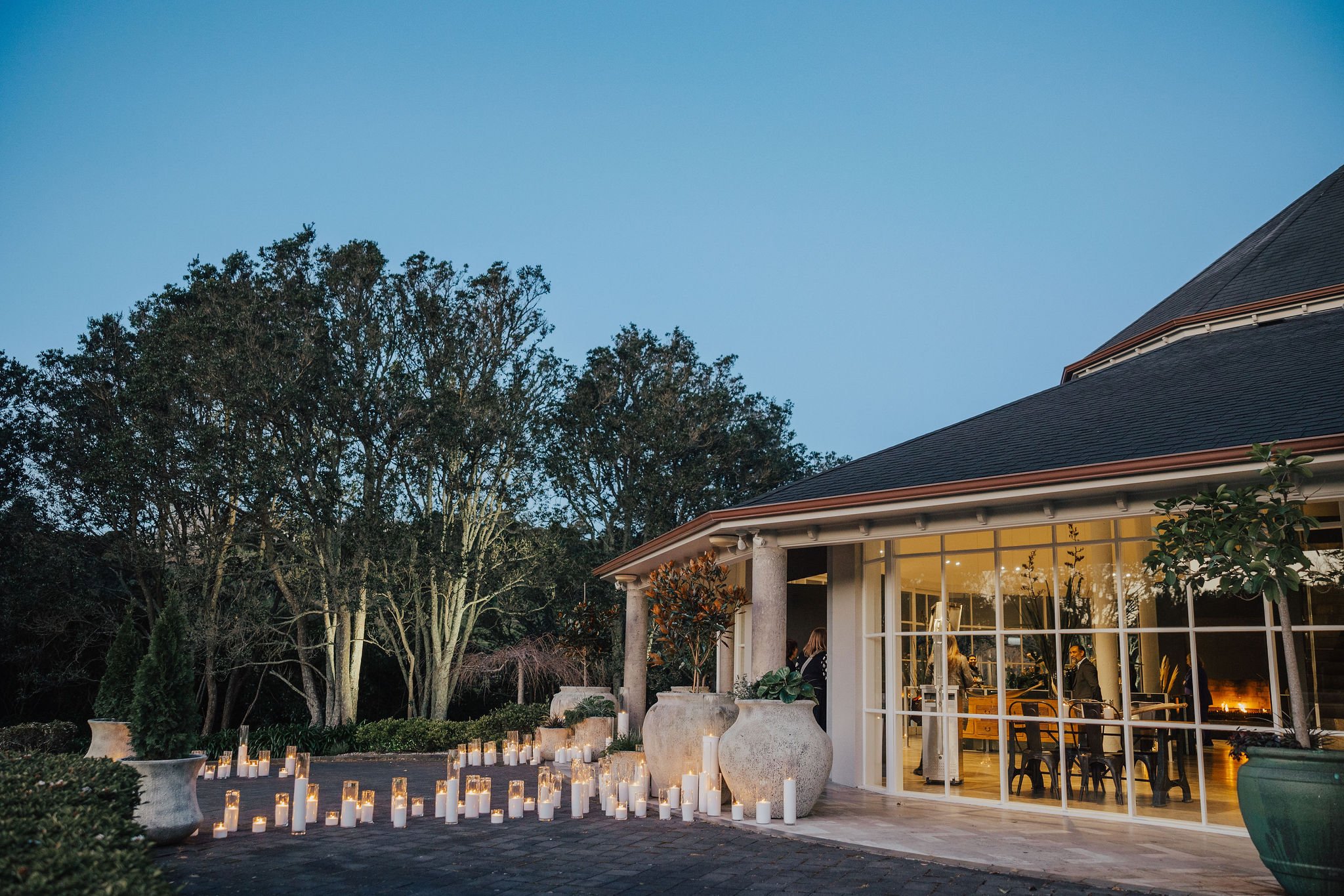 Low-lit exterior at dusk with rows of candles on a paved terrace, glazed pavilion and trees in background
