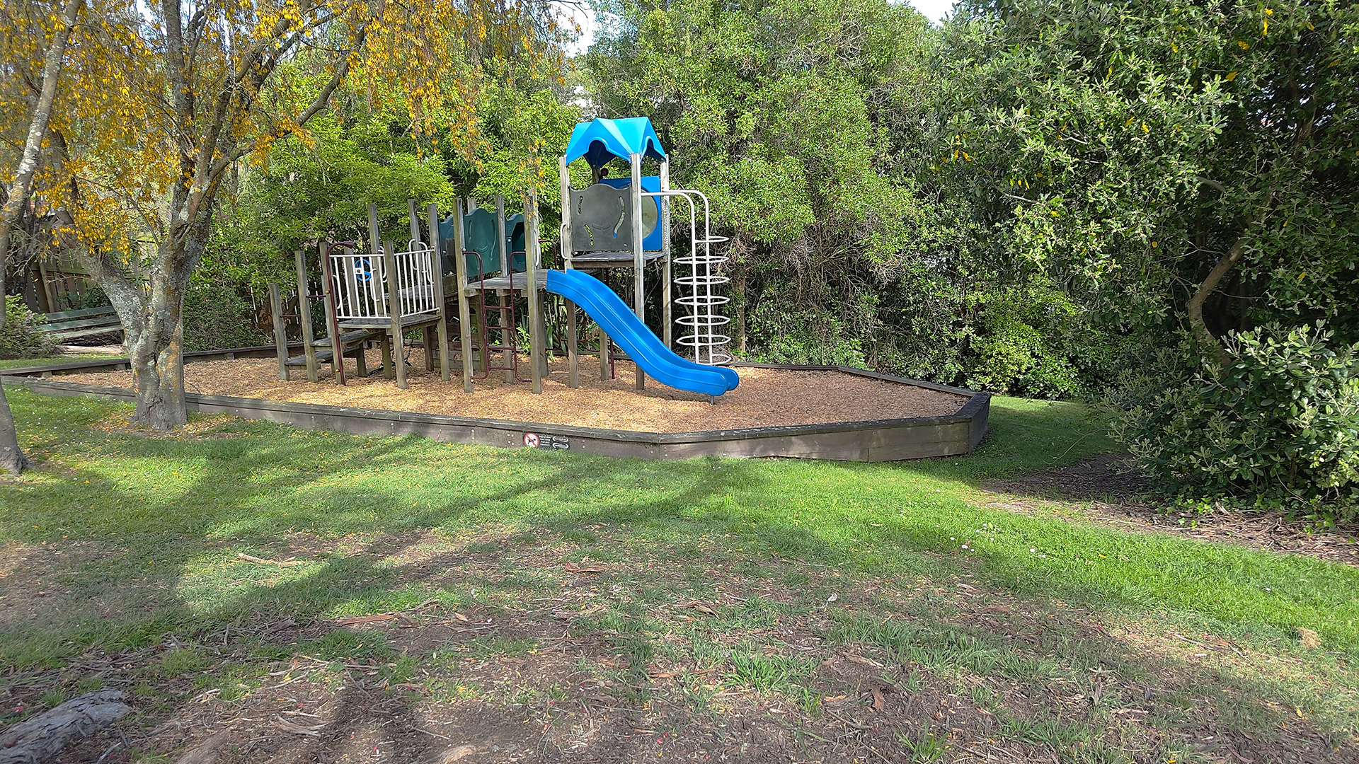 Playground structure with slide surrounded by wood chip and lawn, set among trees