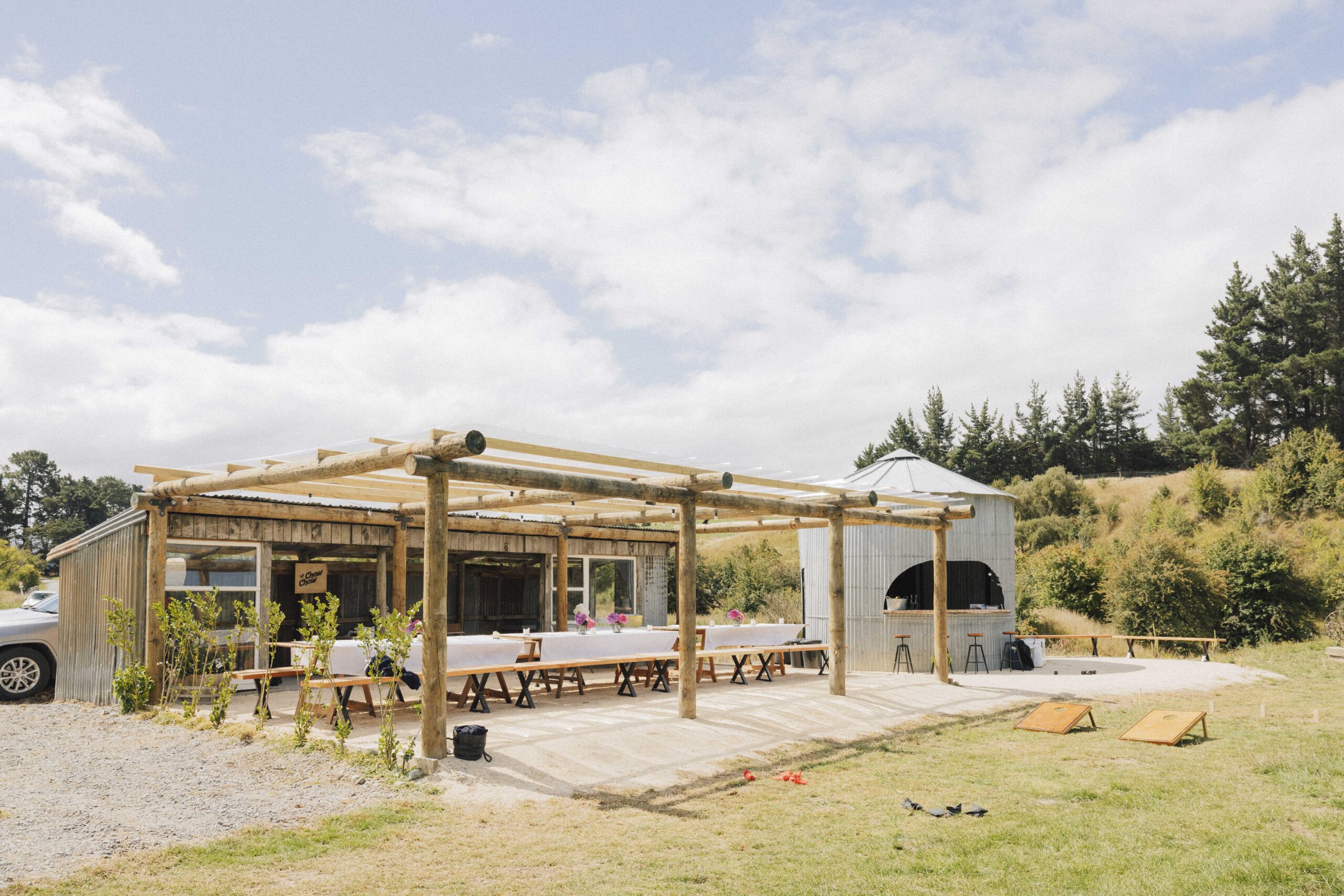 Open timber-frame outdoor shelter with long trestle tables, corrugated iron bar structure beside it, paddock setting