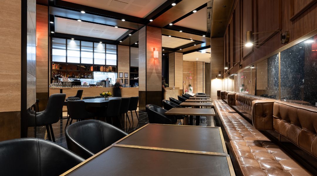 Dark dining room interior with long leather banquette, round tables, warm wall sconces, bar in background