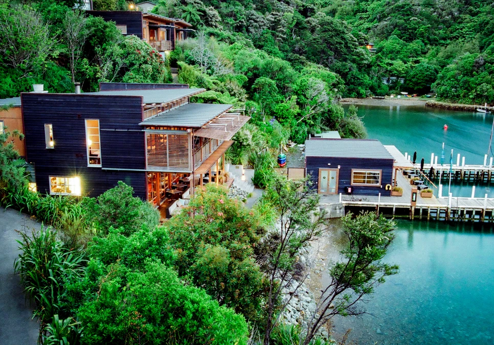 Aerial view of dark-clad lodge buildings set into a steep bush-covered hillside beside a cove