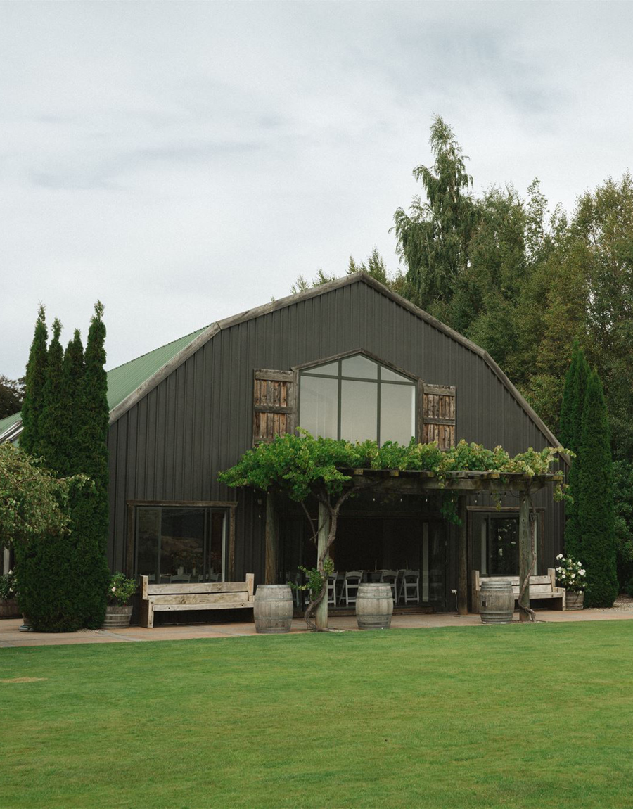 Dark-clad barn venue building viewed across lawn with cypress trees flanking the entrance and wine barrels at the porch
