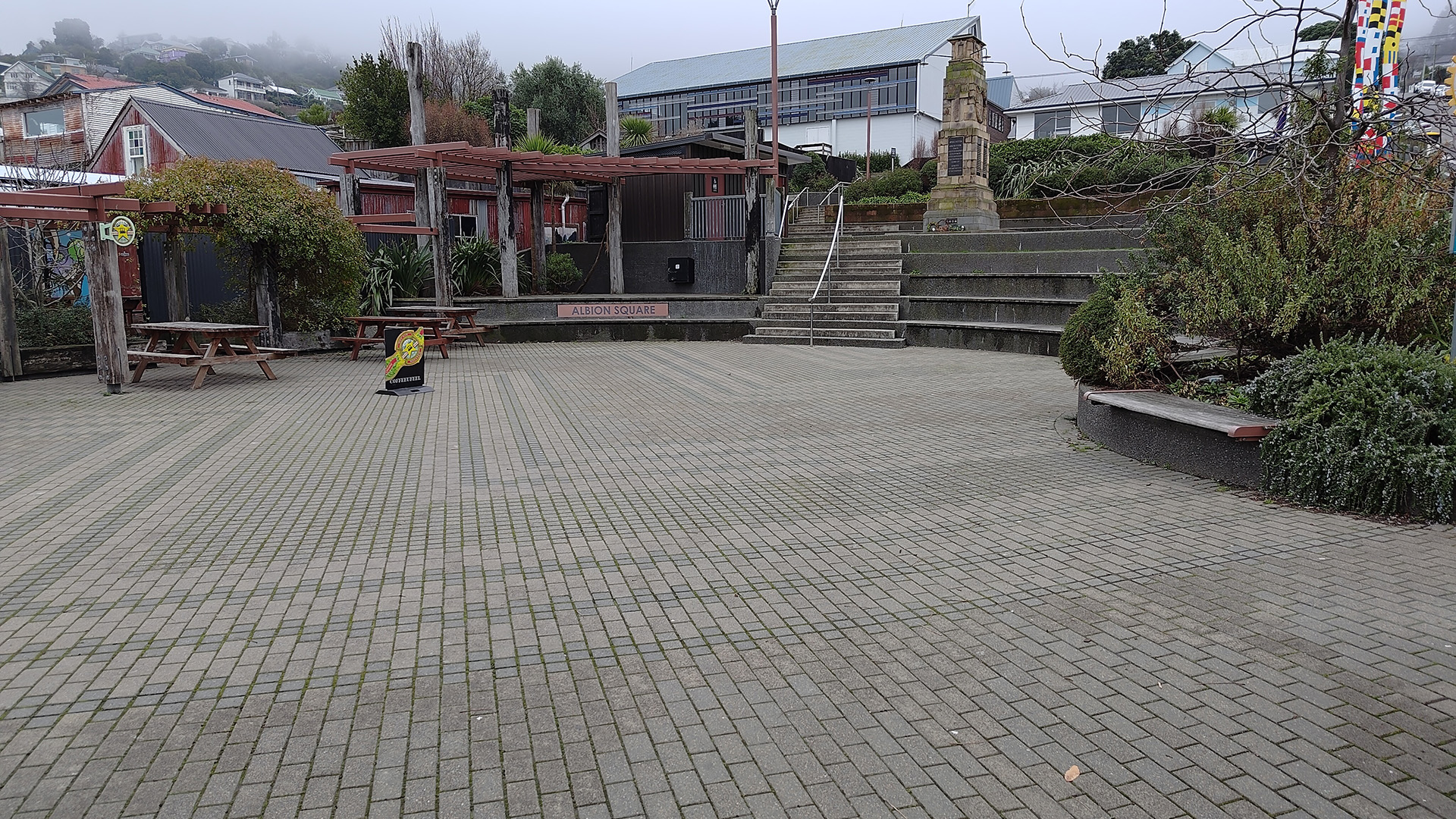 Open paved town square with seating, pergola structures, and memorial column