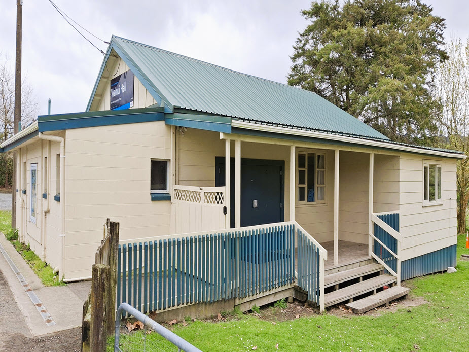 Side view of small weatherboard hall with green roof, veranda and picket fence