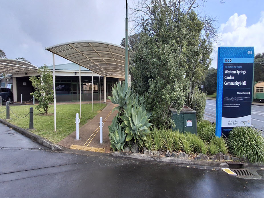 Low-rise hall with curved canopy entrance, sign post, and planted border, roadside setting