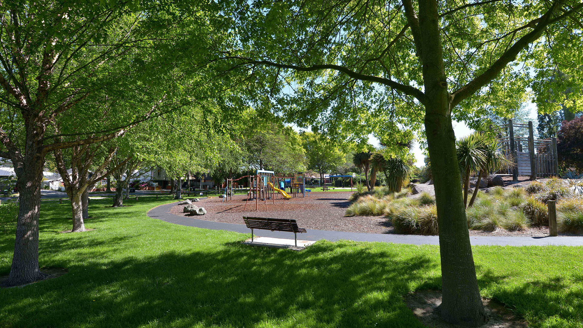 Park with trees, lawn and playground equipment visible, bench in foreground