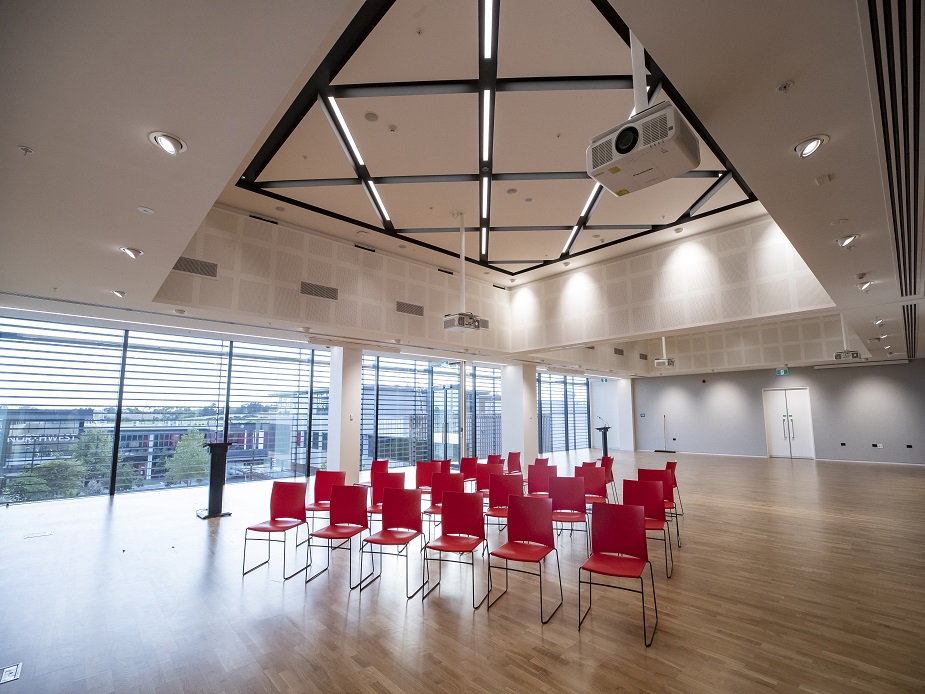 Open hall with timber floor, red chairs in rows, geometric black ceiling frames, and floor-to-ceiling windows