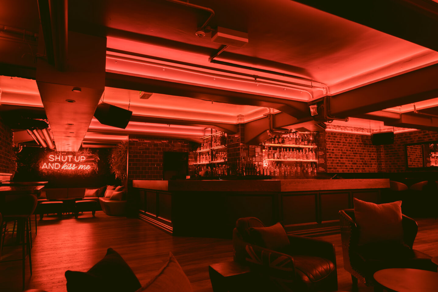 Dark basement bar with red neon lighting, curved bar counter, exposed brick walls, lounge seating