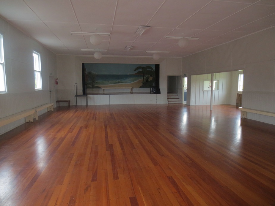 Empty timber-floored hall with polished floor, raised platform at one end with a painted beach mural, mirror on side wall