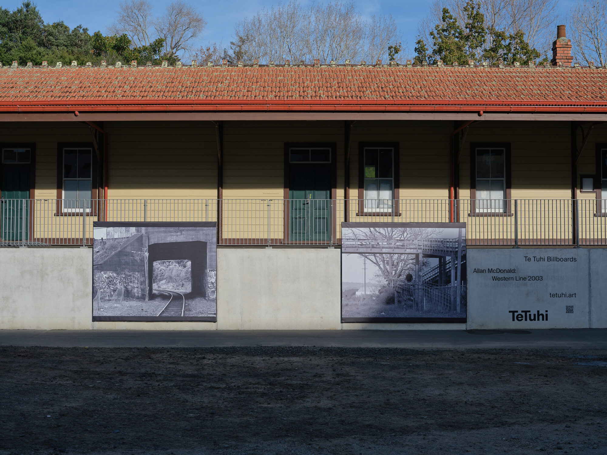 Single-storey yellow weatherboard building with red tile roof, large billboard artworks on base