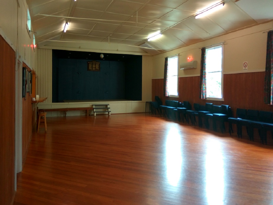Hall interior with polished timber floor, raised platform at far end, and chairs along wall