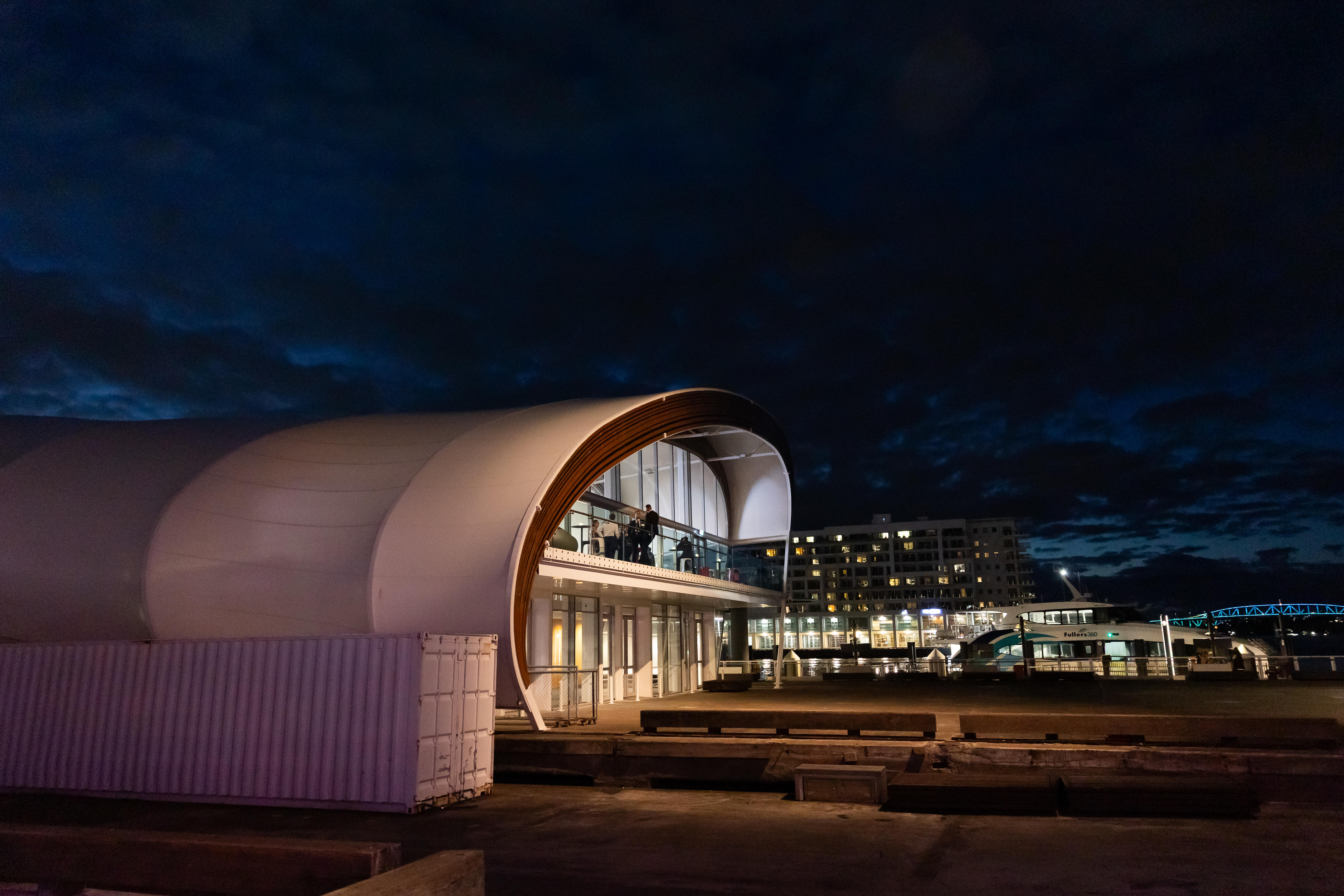 White curved barrel-vault building at night on a waterfront, city lights behind